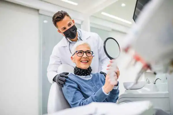 Dentist wearing a face mask standing behind patient sitting in dental chair and looking into handheld mirror